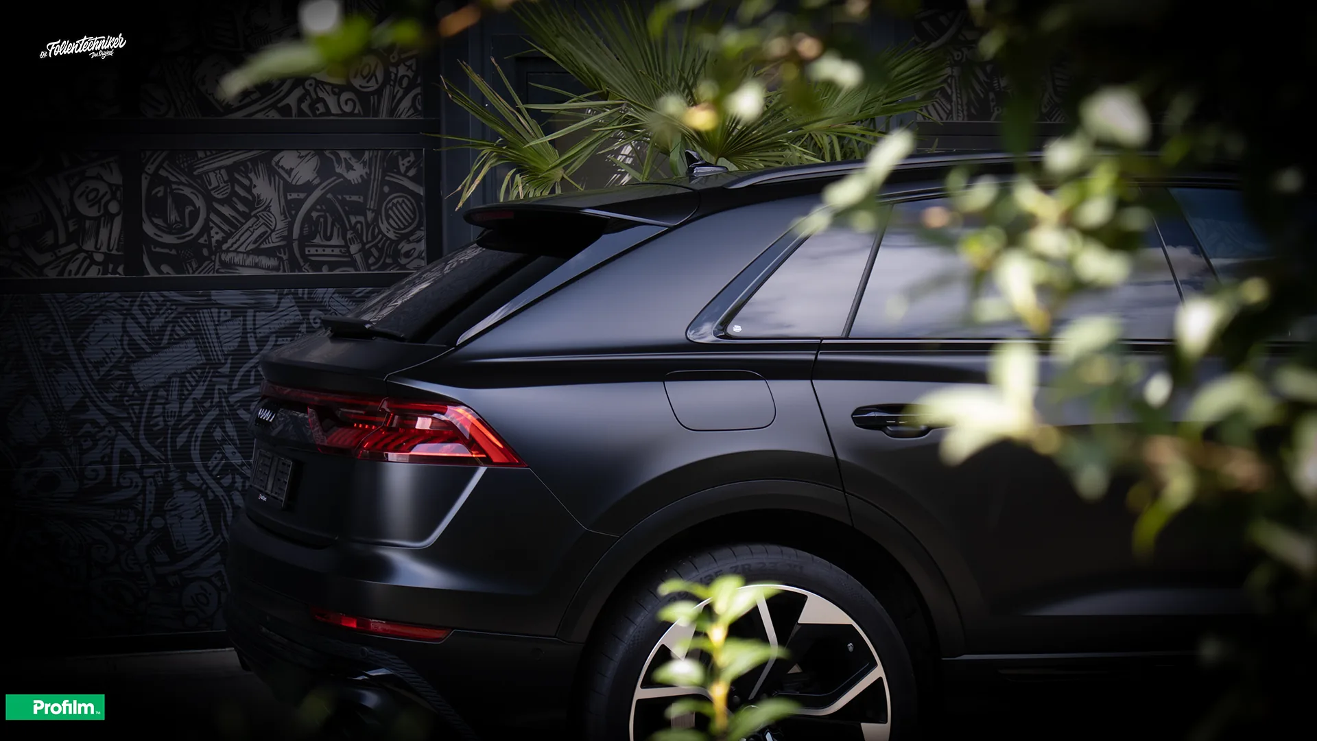 Audi SQ8 in mattem Schwarz, Heckpartie schräg von hinten vor einer dunklen Wand mit Graffiti-Muster
